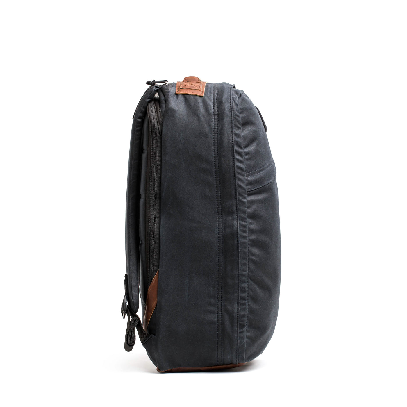The side view showcases the GR1 USA - Heritage backpack by GORUCK, exhibiting a sleek, dark gray design with leather accents and a minimalist aesthetic made from waxed duck canvas. It includes multiple zippers, a top handle, and adjustable shoulder straps. The backpack is photographed against a plain white background.