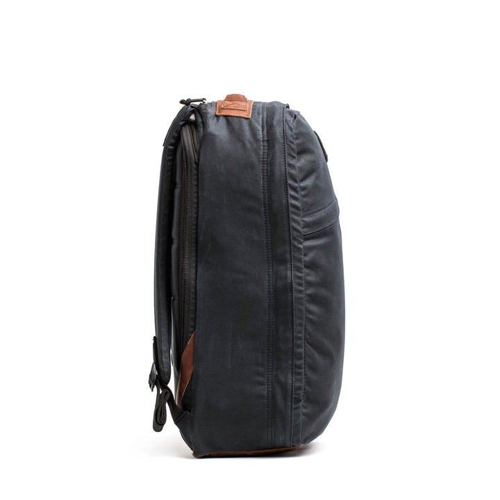 The side view showcases the GR1 USA - Heritage backpack by GORUCK, exhibiting a sleek, dark gray design with leather accents and a minimalist aesthetic made from waxed duck canvas. It includes multiple zippers, a top handle, and adjustable shoulder straps. The backpack is photographed against a plain white background.