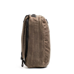 A medium-sized, brown GR1 USA - Heritage rucksack by GORUCK with a top handle and multiple zippers stands on its side against a white background. Crafted from durable fabric, the design features a slim profile with subtle stitching, exuding timeless style.