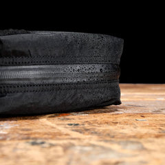 Close-up of the GR2 Field Pocket - X-PAC, a black electronics organizer with AquaGuard zippers, resting on a wooden surface; water droplets showcase its durable, waterproof X-PAC fabric.