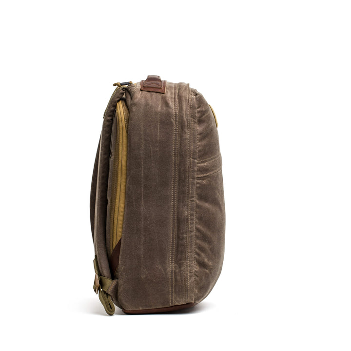 Side view of the GR1 USA - Heritage by GORUCK, a brown waxed canvas rucksack featuring a visible zipper and leather accents, standing upright against a plain white backdrop.