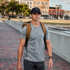 Man in a durable Slick Short Sleeve Tee - Tri-BlendX gray shirt, black cap, and brown backpack walking on a brick path near buildings and grass.