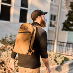 Man wearing a tan GORUCK backpack walking outdoors near a building in casual rucking gear