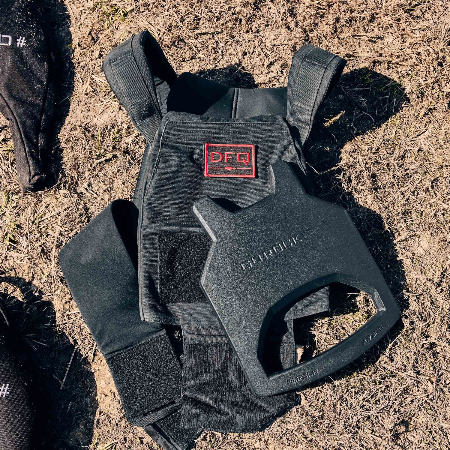 A black rucking weight vest and a Curved Ruck Plate rest on dry grass outdoors.