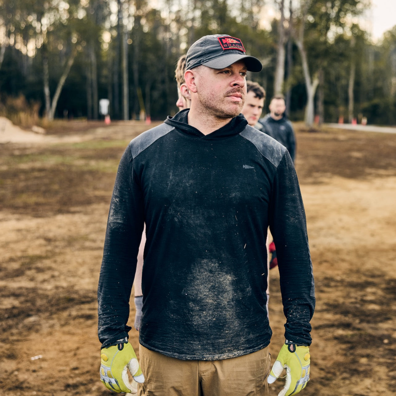 A man wearing the Men’s Commando Pullover - Merino Wool and gloves stands outdoors on dirt, with three people blurred in the background.