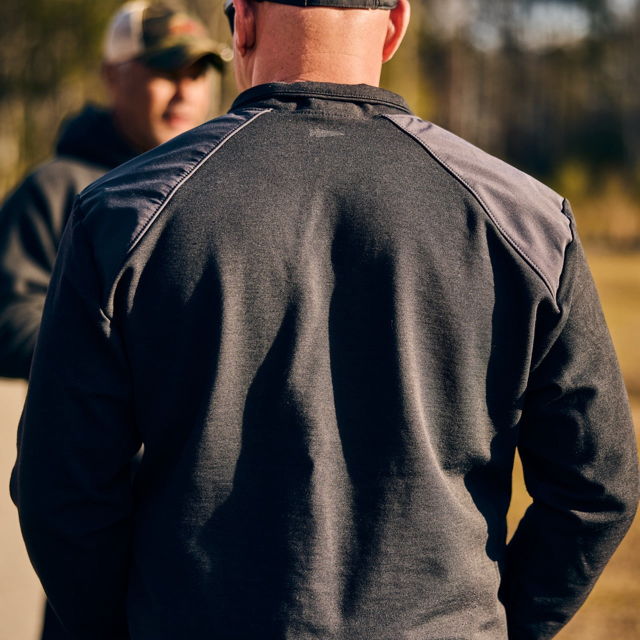 A man wearing the Men's Commando Quarter Zip - Merino Wool stands outdoors with his back to the camera; another person is blurred in the background.