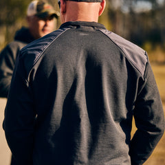 A man wearing the Men's Commando Quarter Zip - Merino Wool stands outdoors with his back to the camera; another person is blurred in the background.