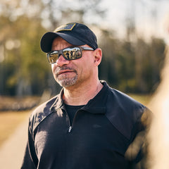 A man wears the Men’s Commando Quarter Zip—crafted from quick-drying, antimicrobial Merino wool—with a black cap and sunglasses, standing outdoors amid trees and sunlight.