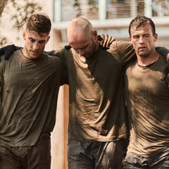 Three men in muddy Slick Short Sleeve Tee - Tri-BlendX shirts support each other, arms over shoulders, looking tired and serious outdoors.