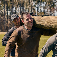 Two men outdoors, both wearing the Men’s Commando Long Sleeve - Merino Wool shirts with reinforced shoulders, team up to carry a large wooden log during a physical challenge.