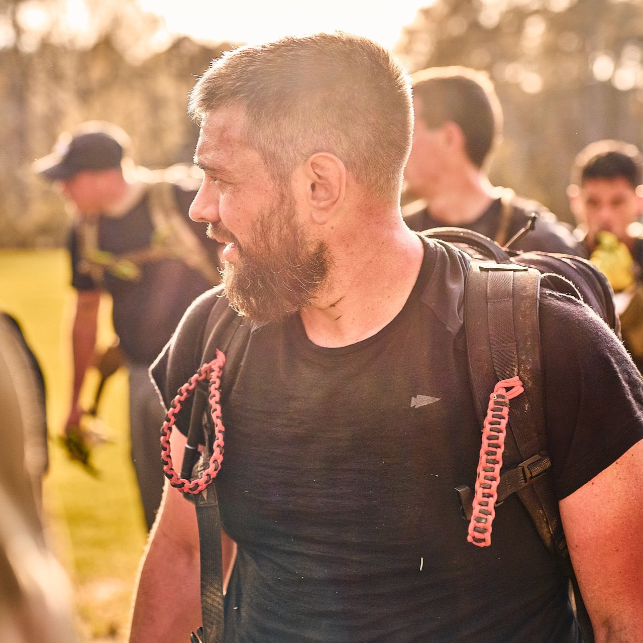 A bearded man wearing the Men’s Commando Tee - Merino Wool with reinforced shoulders and a backpack stands outdoors with others in warm sunlight.