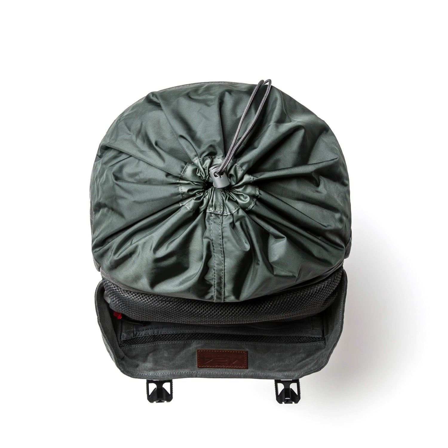 A green M24 rucksack from GORUCK, featuring a drawstring closure on top, is open to display its spacious interior. Made from robust waxed canvas, two small straps dangle from the front flap, and it is set against a plain white background for the photograph.
