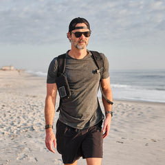 Man in sunglasses, cap, and backpack walking on a sandy beach with the ocean and sky in the background, wearing the Slick Short Sleeve Tee - Tri-BlendX for ultimate comfort and freshness.