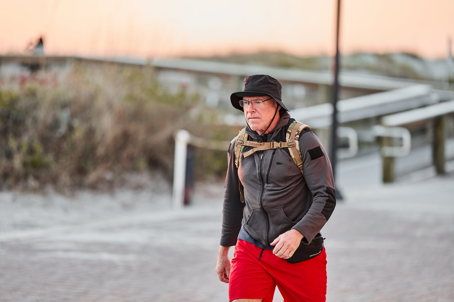A man wearing the RPC 3.0 + 10lb Ruck Sandplate walks on a paved beachside path at sunset, dressed in a black hat, gray jacket, and red shorts.