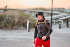 A man wearing the RPC 3.0 + 10lb Ruck Sandplate walks on a paved beachside path at sunset, dressed in a black hat, gray jacket, and red shorts.