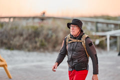 An older man in a hat and backpack walks outdoors by the beach at sunset, training with his RPC 3.0 + 10lb Ruck Sandplate.