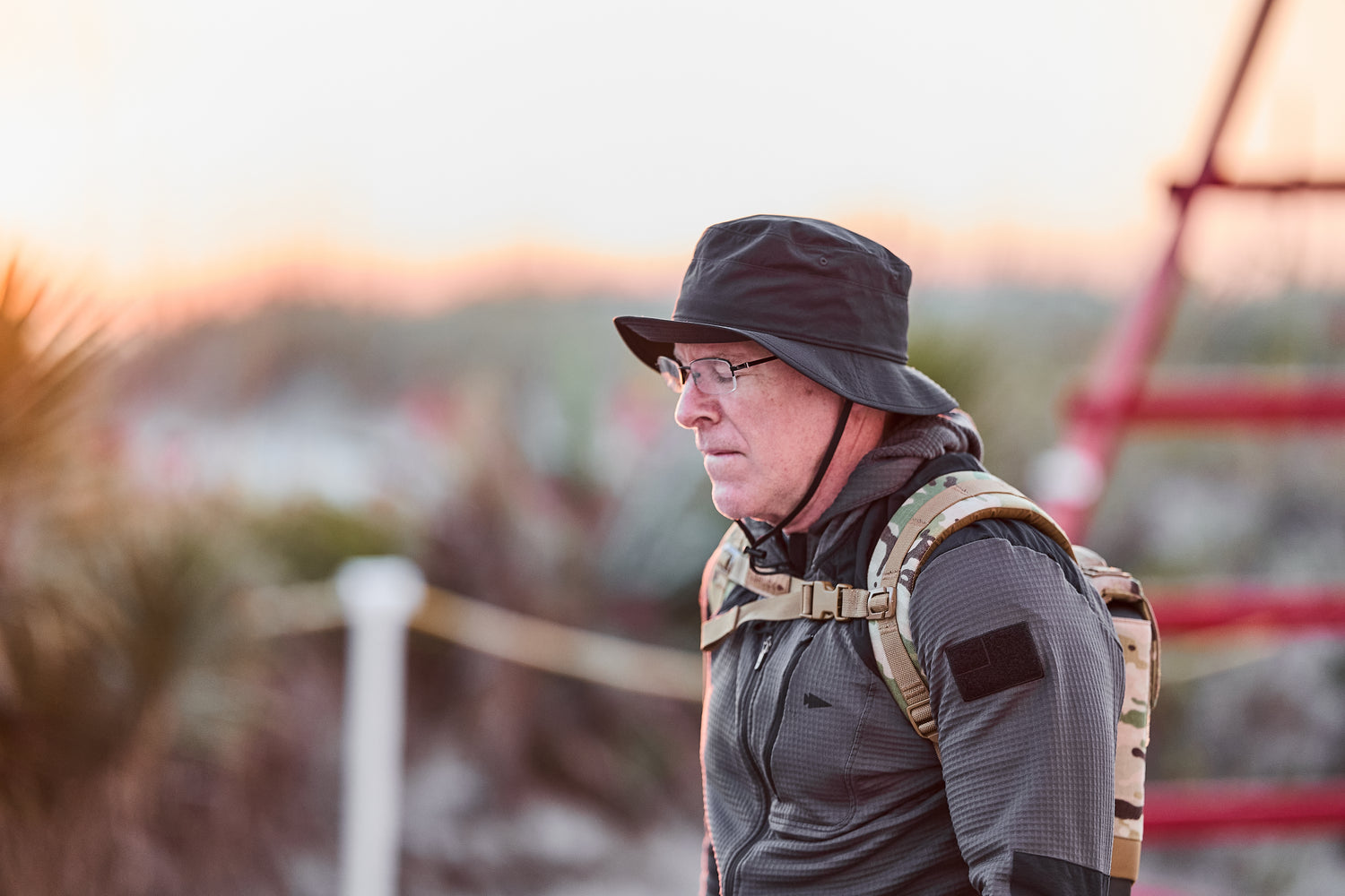 A man wearing a black hat and the RPC 3.0 + 10lb Ruck Sandplate walks outdoors, with greenery and a red structure blurred in the background.