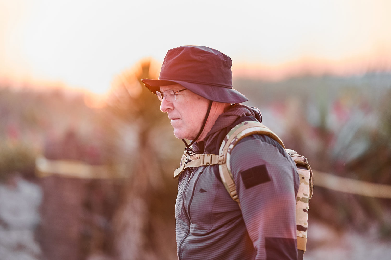 A man wearing glasses, a black hat, and a backpack (RPC 3.0 + 10lb Ruck Sandplate) walks outdoors at sunset.