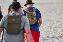 Two people with backpacks and hats, likely using the RPC 3.0 + 10lb Ruck Sandplate for weighted training, walk away from the camera on a sandy surface.