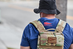 A person in a blue shirt and black hat wears the RPC 3.0 + 10lb Ruck Sandplate for weighted training while walking outdoors.