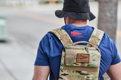 Person walking outdoors in a blue GORUCK shirt and camouflage vest, wearing a black bucket hat and carrying the RPC 3.0 + 10lb Ruck Sandplate.