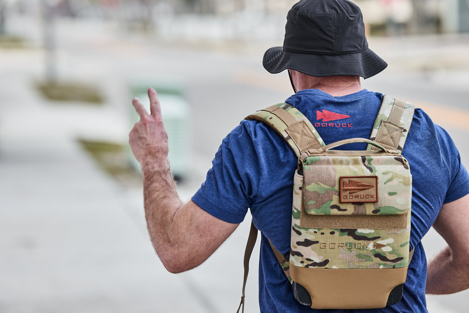 Man wearing a bucket hat, blue GORUCK shirt, and carrying an RPC 3.0 + 10lb Ruck Sandplate backpack flashes a peace sign while walking outside.