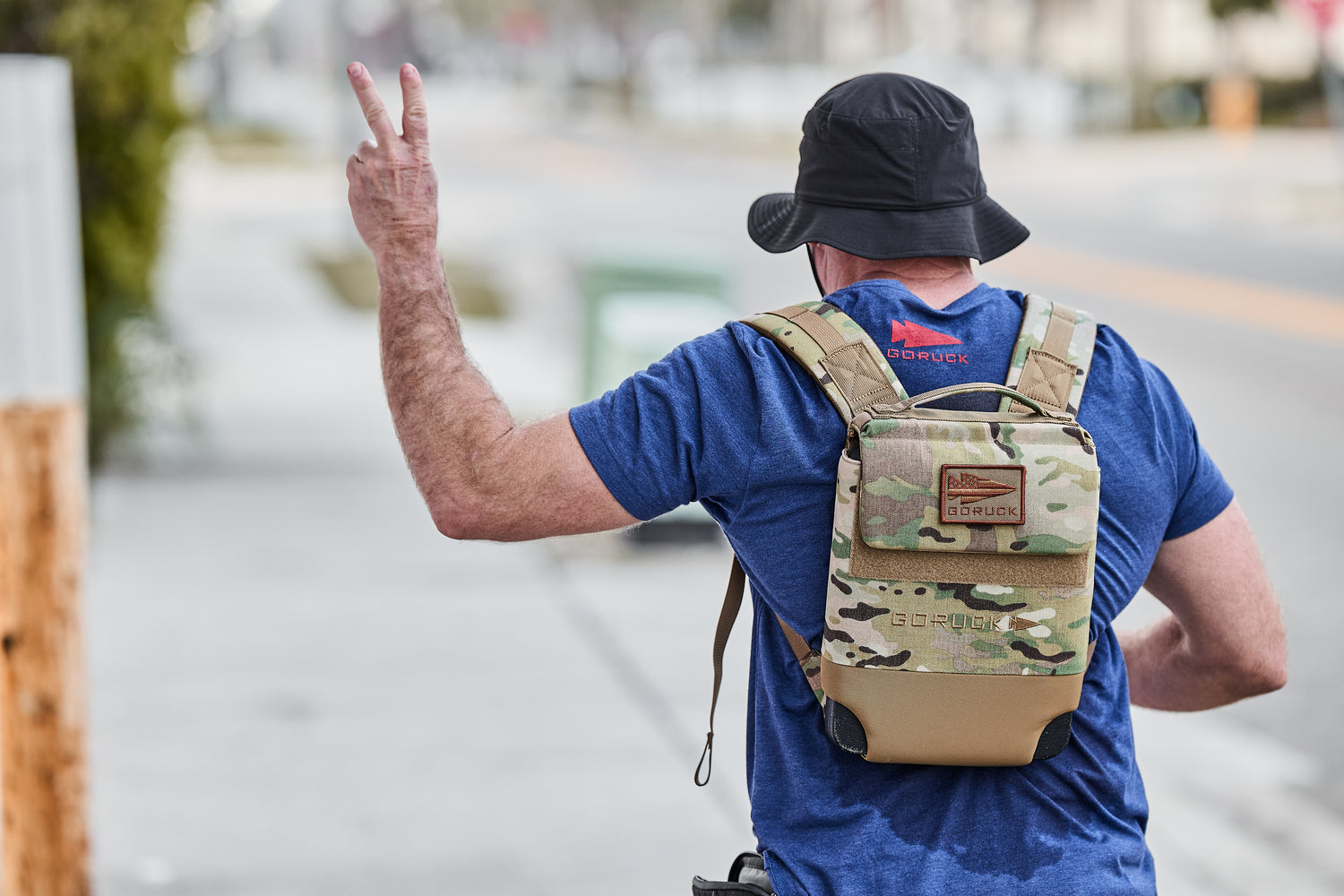 Man wearing a bucket hat and carrying the RPC 3.0 + 10lb Ruck Sandplate backpack flashes a peace sign while walking down the street.