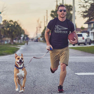 Wearing Men’s Simple Shorts - Lightweight ToughDry® and sunglasses, a man jogs with his dog on a leash, expertly holding a football. These 2-way stretch, fast-drying shorts effortlessly match his lively outdoor adventures.