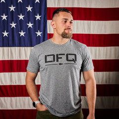 A man wearing the DFQ Tee - Tri-BlendX, a gray premium blend t-shirt with "DFQ" on the front, stands before a large American flag and gazes to the side.