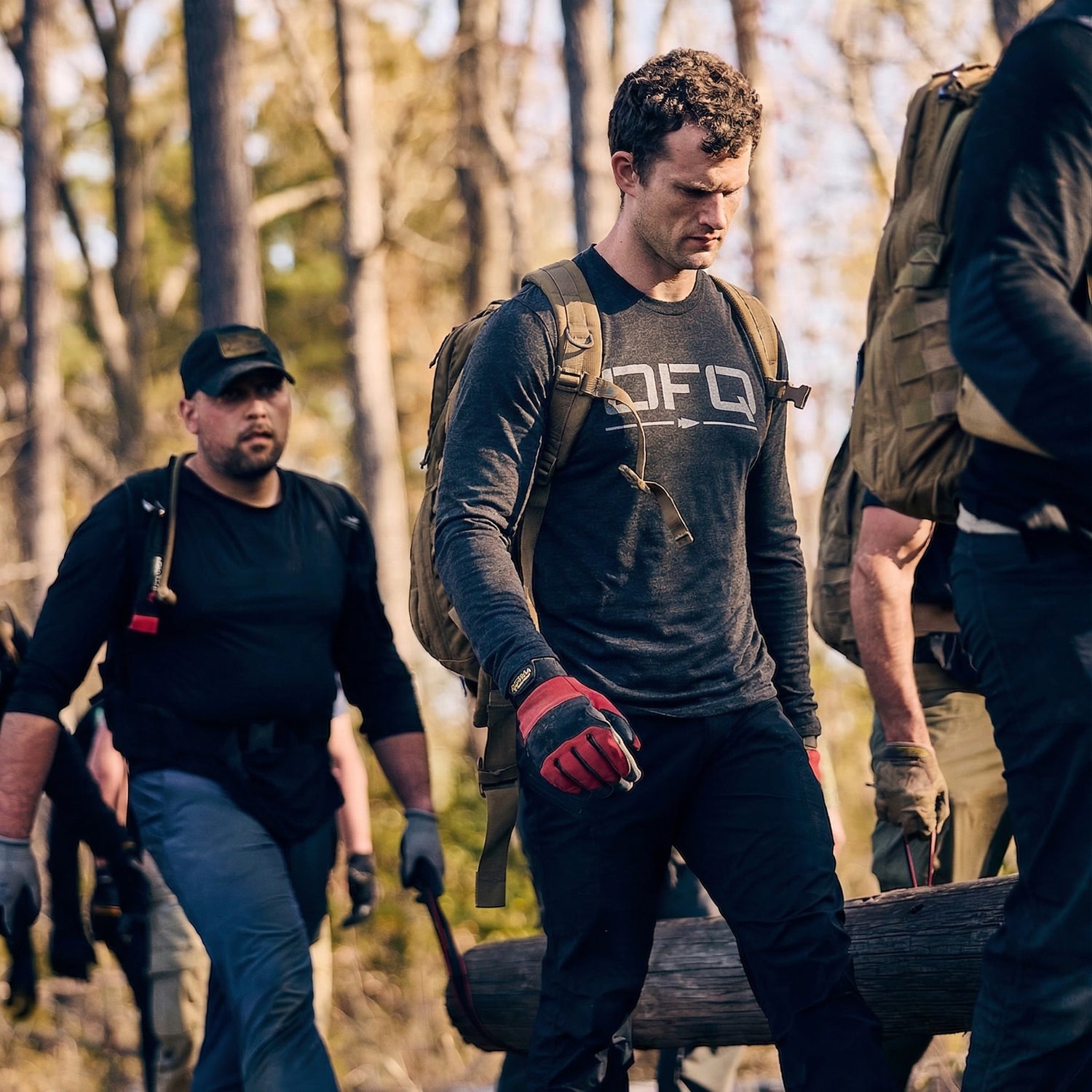 A group in outdoor gear, all wearing Men's DFQ Long Sleeve - Tri-BlendX shirts with antimicrobial treatment, carry a heavy log together through a forested area.
