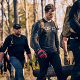 A group in outdoor gear, all wearing Men's DFQ Long Sleeve - Tri-BlendX shirts with antimicrobial treatment, carry a heavy log together through a forested area.