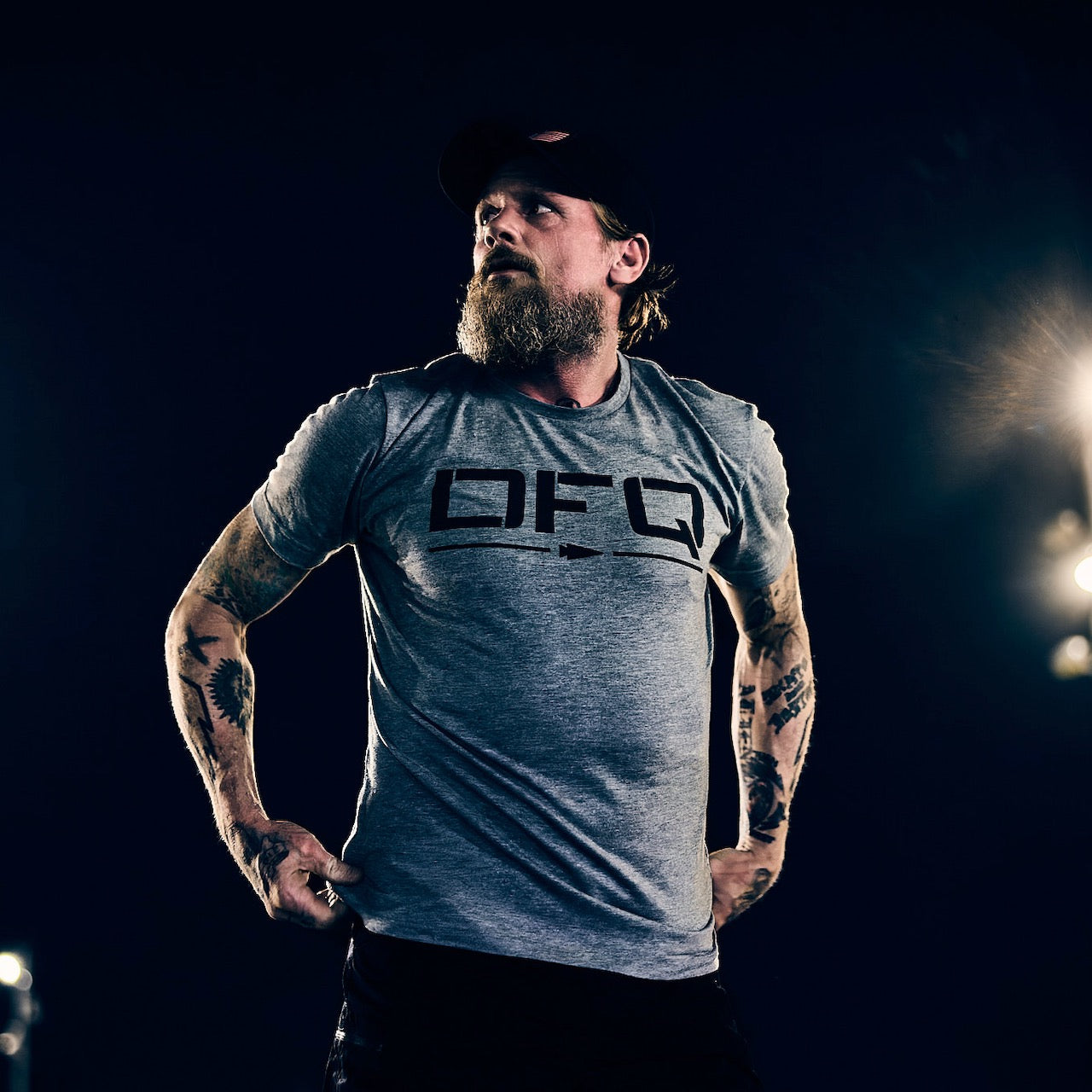 A bearded man in a gray DFQ Tee - Tri-BlendX stands confidently, gazing to the side under dramatic lighting. Made from our premium Tri-BlendX fabric, this shirt delivers unmatched comfort and bold attitude for those who never back down.