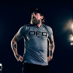 A bearded man in a gray DFQ Tee - Tri-BlendX stands confidently, gazing to the side under dramatic lighting. Made from our premium Tri-BlendX fabric, this shirt delivers unmatched comfort and bold attitude for those who never back down.
