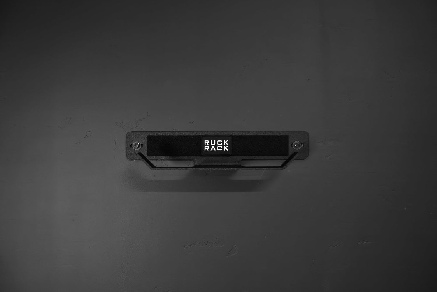 A Ruck Rack® Delta, a heavy-duty black wall-mounted storage rack for rucks, weight vests, and tactical gear, is centered on a dark wall, serving both as a versatile organizer and practical display solution.