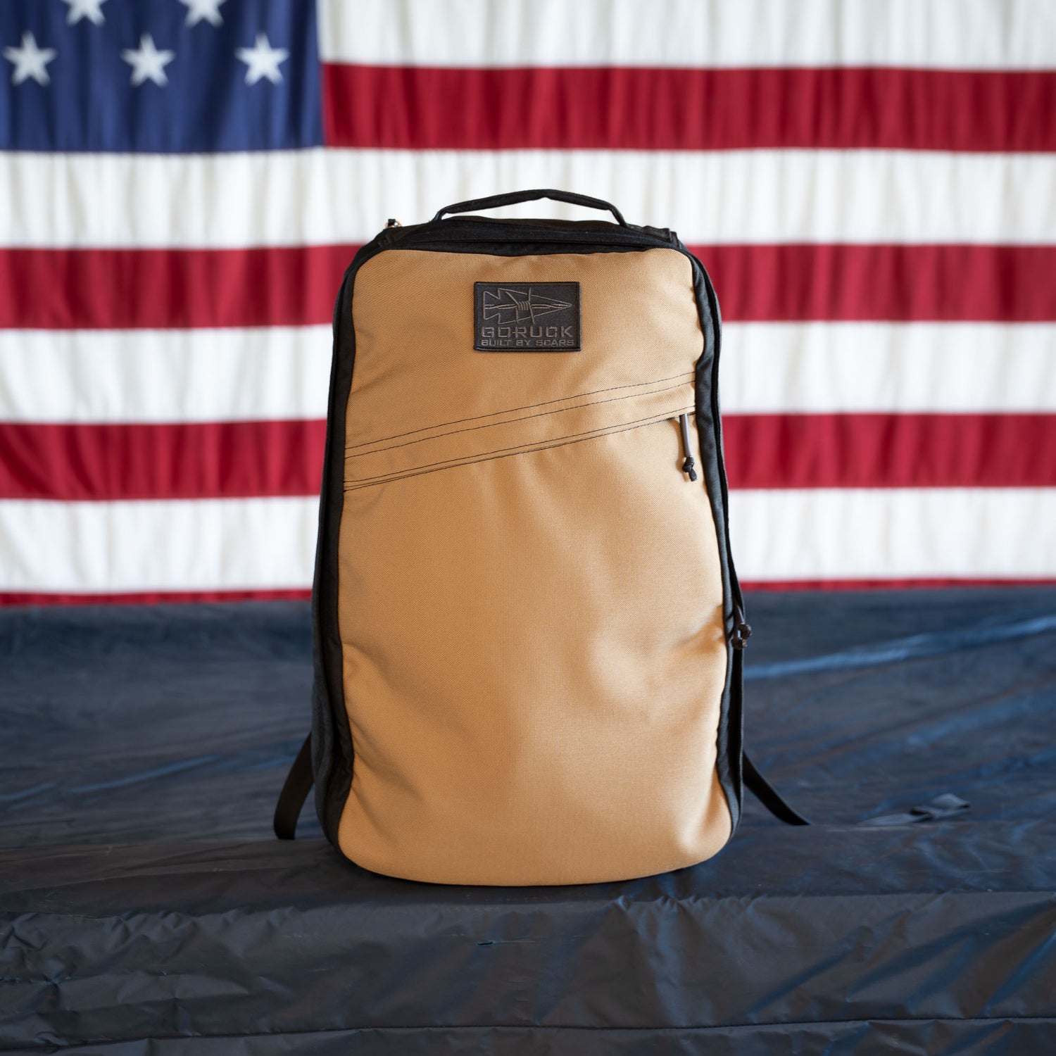 A tan GR1 Shooter - Built by SCARS backpack rests on a dark surface with a large American flag behind it, representing rugged gear trusted by the SCARS team.