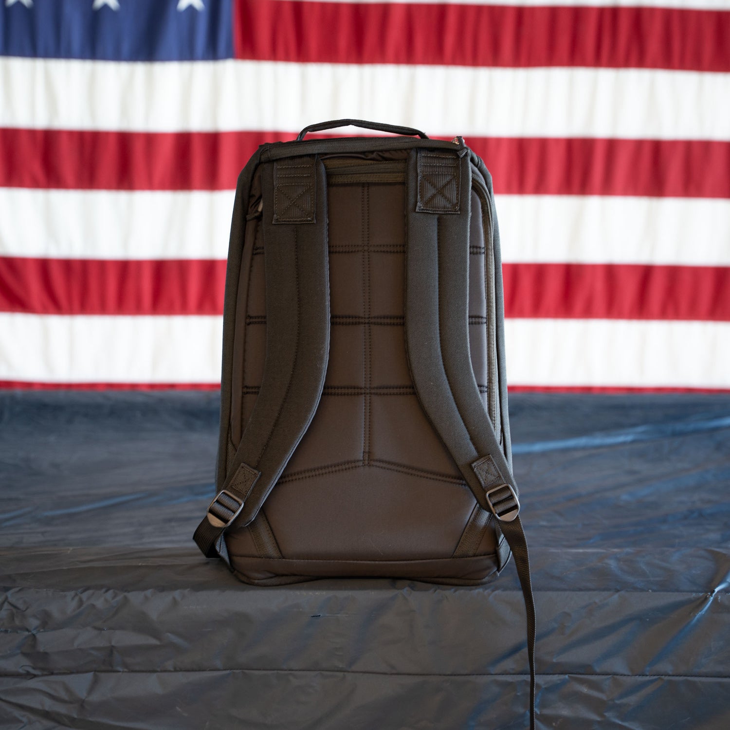 A brown GR1 Shooter - Built by Scars backpack sits on a gray surface with an American flag in the background, reflecting the SCARS team spirit.