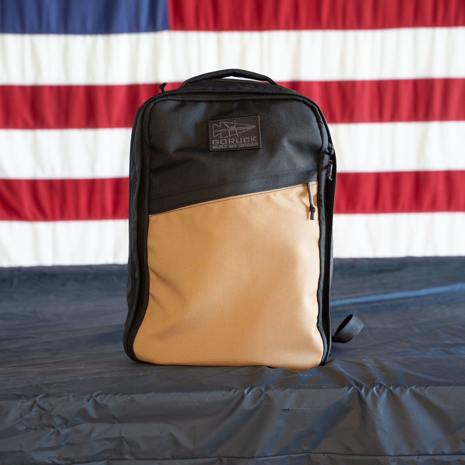A tan and black GR1 Shooter - Built by scars backpack sits on a table with an American flag in the background.