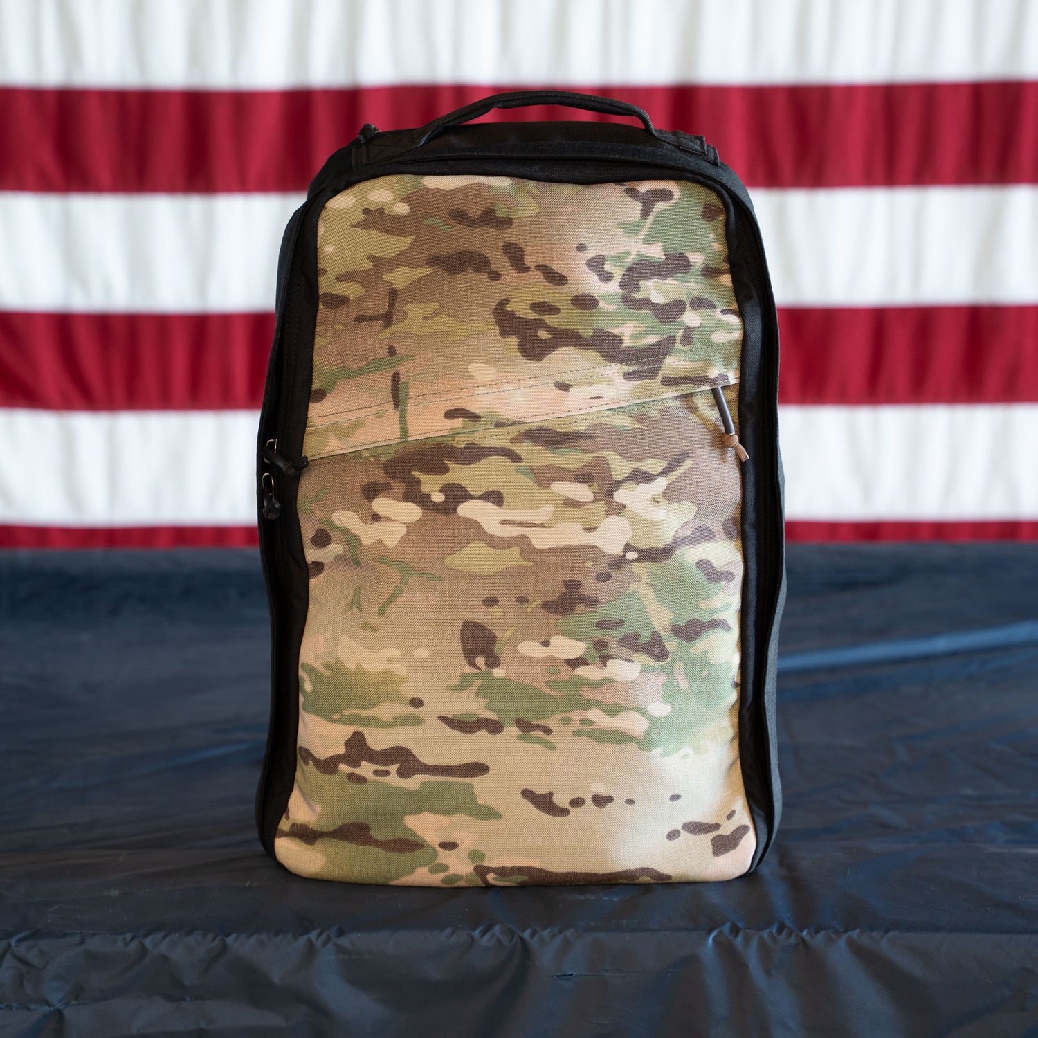 A camouflage GR1 Shooter - Built by SCARS backpack rests on a dark surface with an American flag behind it, showcasing the rugged durability trusted by the SCARS team.