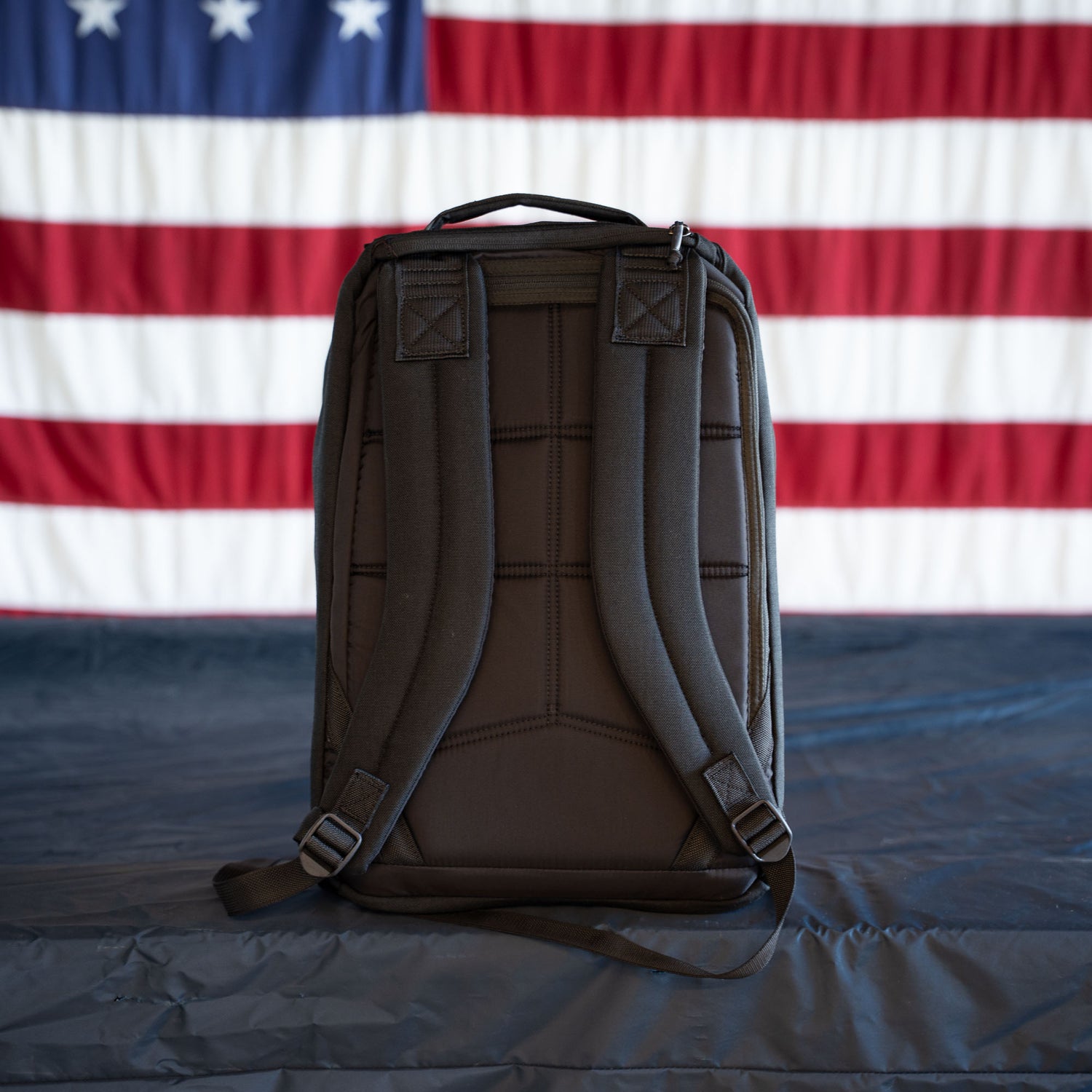 A black GR1 Shooter - Built by Scars ruck stands upright on a dark surface with a U.S. flag hanging in the background.