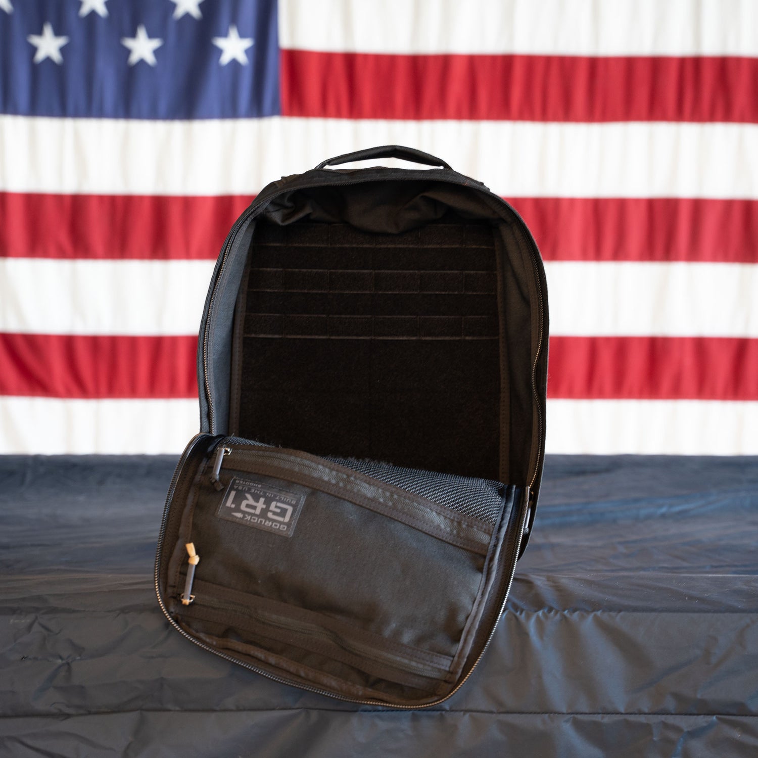 An open black GR1 Shooter - Built by Scars backpack rests on a table, with an American flag displayed in the background.