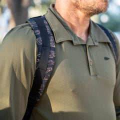 Man outdoors, ready for rucking, wears an olive green collared shirt and carries the Basic Rucker® backpack featuring black and patterned straps.