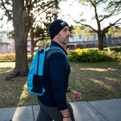 A man rucks outdoors in a sunlit park, wearing a black beanie and carrying the blue Basic Rucker® backpack—possibly loaded with a ruck plate—surrounded by trees.