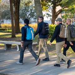 Four men carrying Basic Rucker® backpacks walk together on a sunny park sidewalk lined with trees and grass.