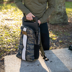 A person slides a GORUCK ruck plate into a Basic Rucker® camouflage backpack outside on a sunny day, prepared for serious rucking.