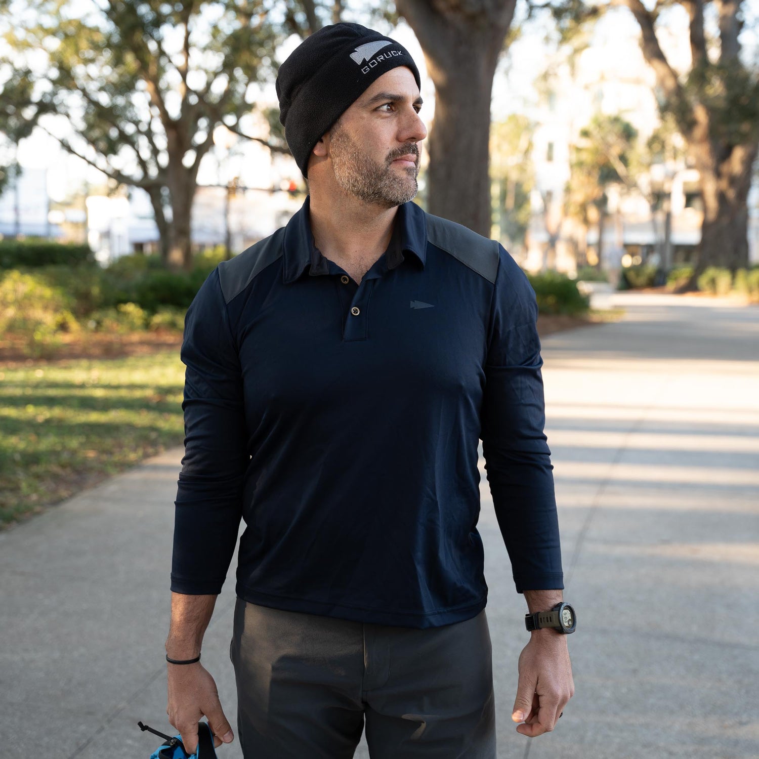 A man stands outdoors on a sunny day, wearing the Men's Long Sleeve Rucking Polo and a black beanie, looking to the side.