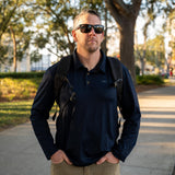 A man stands outdoors on a sunny day, wearing sunglasses, a backpack, and the Men's Long Sleeve Rucking Polo made in the USA, with trees in the background.
