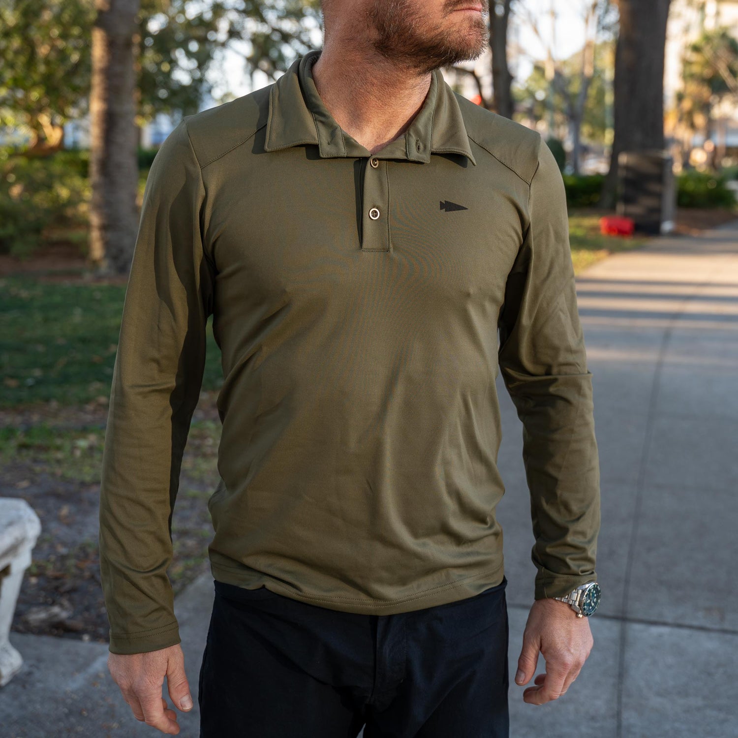 A man stands outdoors on a park sidewalk wearing the Men's Long Sleeve Rucking Polo in olive green, made with durable EnduraKnit fabric.