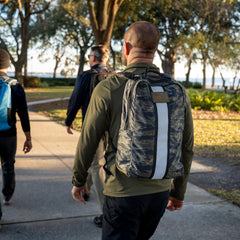 Three people with backpacks and ruck plates walk on a sunlit park path lined with trees and green grass, enjoying a rucking session with the Basic Rucker®.
