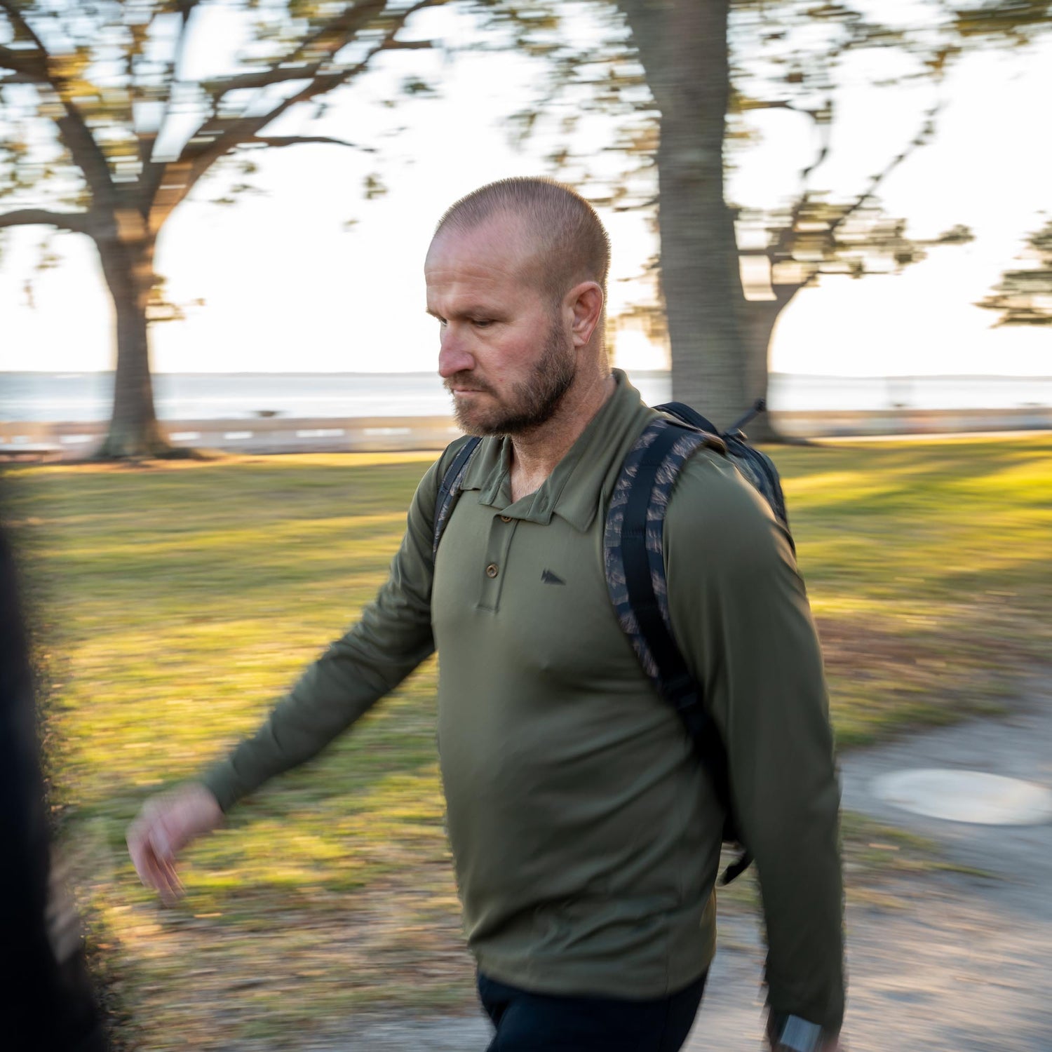 A bearded man with a backpack walks in a sunlit park, wearing the Men's Long Sleeve Rucking Polo, made in the USA.