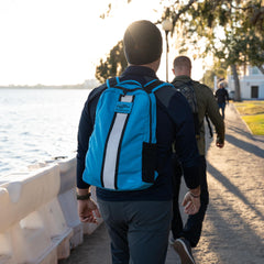 A person wearing a blue Basic Rucker® backpack walks with others along a waterfront path on a sunny day.